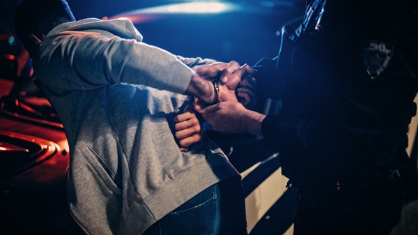 Police officer handcuffing a man next to a patrol car at night with flashing lights.