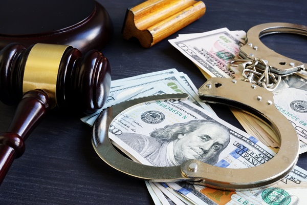 A judge's gavel rests on a dark surface next to a pair of open handcuffs and scattered stacks of $100 and $50 bills.