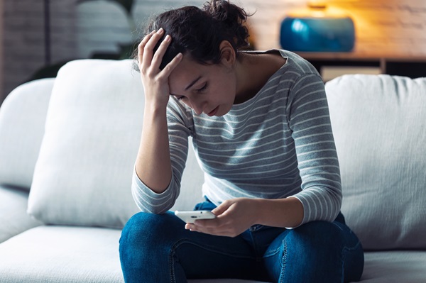 A woman sitting on a couch, looking down at a smartphone with her hand on her forehead in a stressed or worried manner.