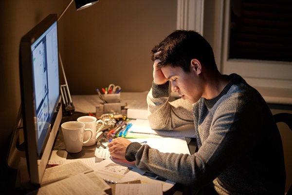 A stressed college student sits at a desk late at night with his head in his hands, reflecting the academic and professional pressure caused by Title IX investigation delays, graduation barriers, and transcript holds.